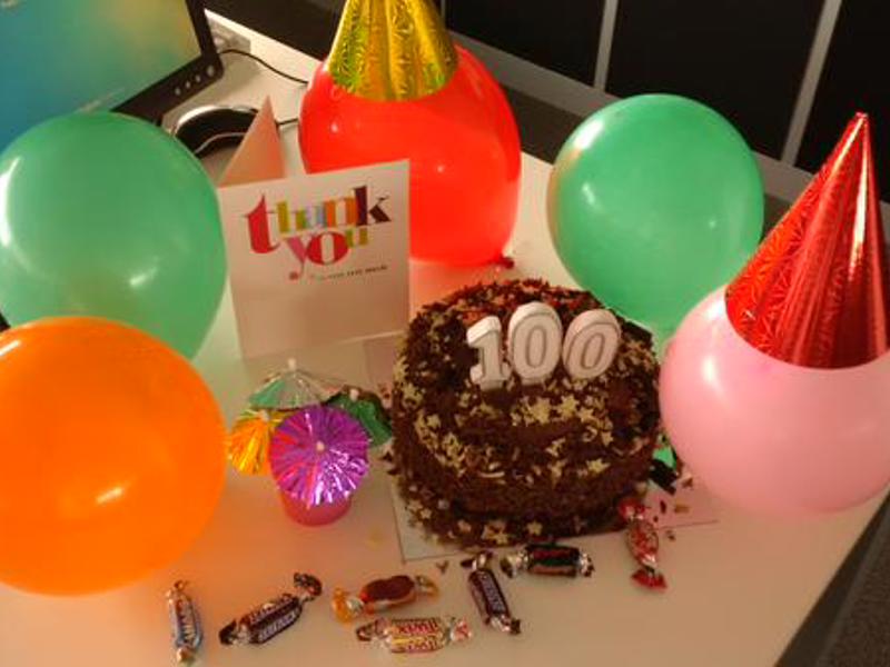 Cake with '100' candle and balloons on a desk.
