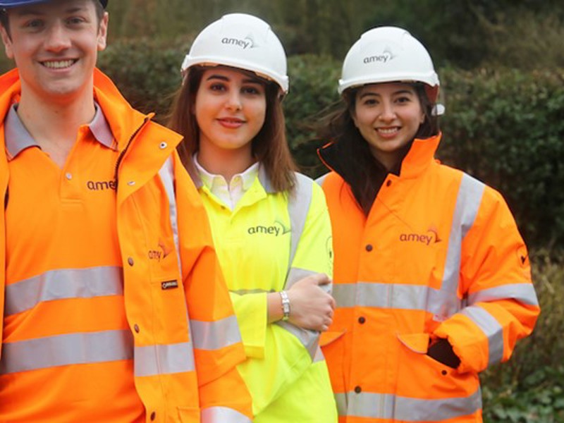 Three young Amey employees, stood one in front of the other, wearing PPE, smiling at the camera.