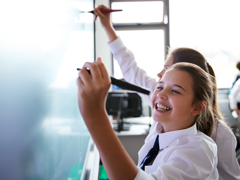 Two school pupils standing side by side, actively writing on a large whiteboard with markers