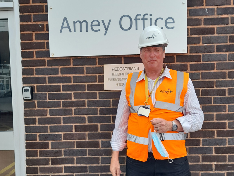 Male Amey employee, wearing a hi vis jacket, stood in front of an Amey office sign.