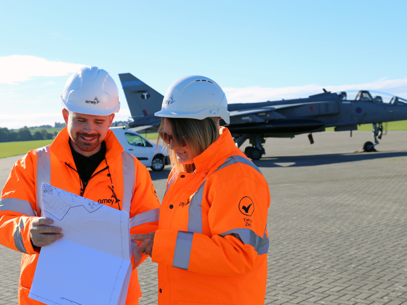 Two Amey employees, wearing PPE, inspecting a map, stood in front of an aircraft.