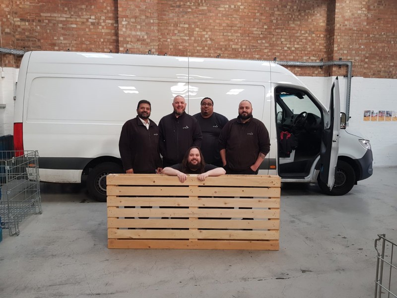 Image of five men, standing holding a palate in front of a van.