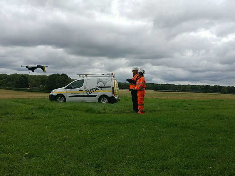 image of a drone in flight in a field, with two Amey employees standing by a van.