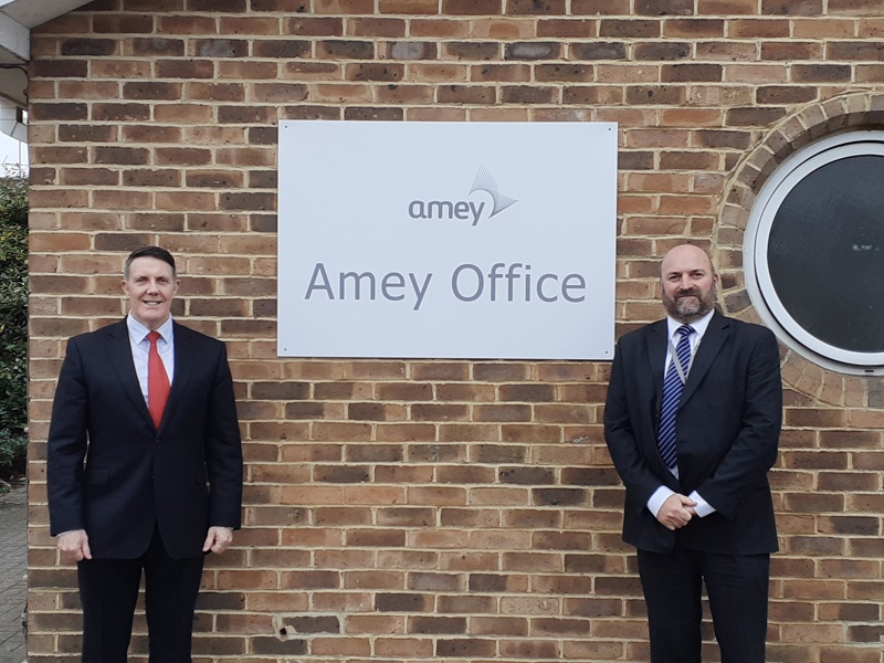 Image of ex lt col who has just joined Amey, stood in front of an Amey office sign with another Amey employee.